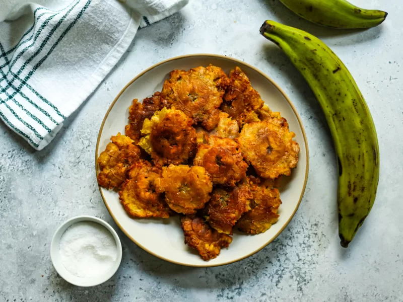 Tostones on a plate with a green plantain nearby